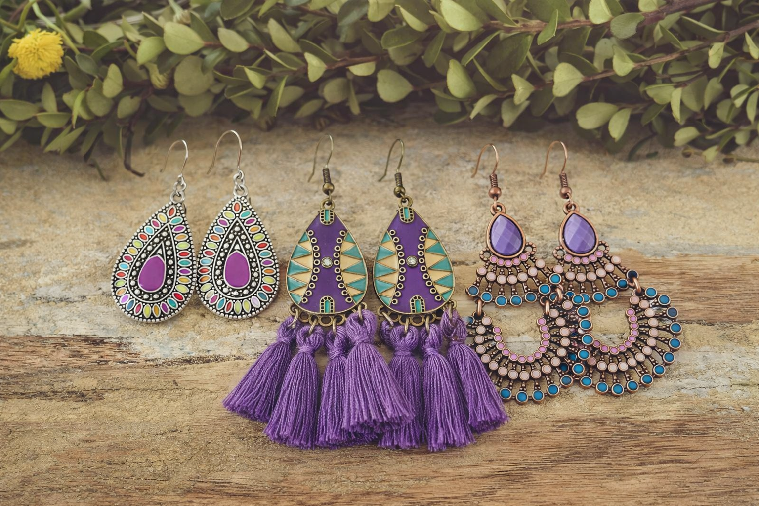 Three pairs of vibrant dangle earrings displayed on a wooden surface with green leaves, featuring colorful enamel teardrops, ornate purple tassel designs, and elegant copper chandelier earrings with purple stones