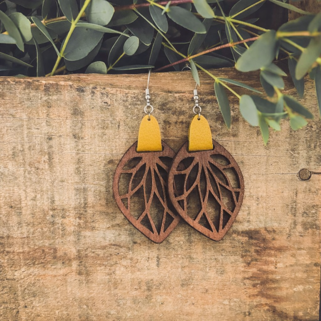 A pair of handmade wooden leaf-cutout earrings with bright yellow leather accents and silver hooks hanging against a rustic wooden board with fresh green eucalyptus leaves overhead