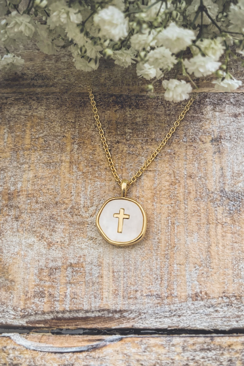 Photograph of a delicate gold chain necklace featuring a wavy white mother-of-pearl pendant with a centered gold cross, resting on a rustic wooden background under a cluster of soft white baby's breath flowers