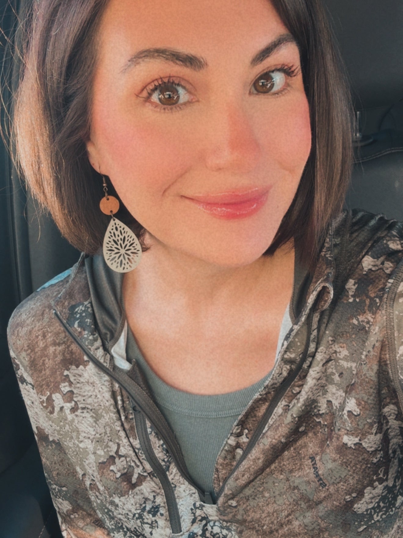 Smiling woman with dark bob hair and natural makeup wearing a wooden and white filigree dangle earring and a camouflage hoodie inside a car