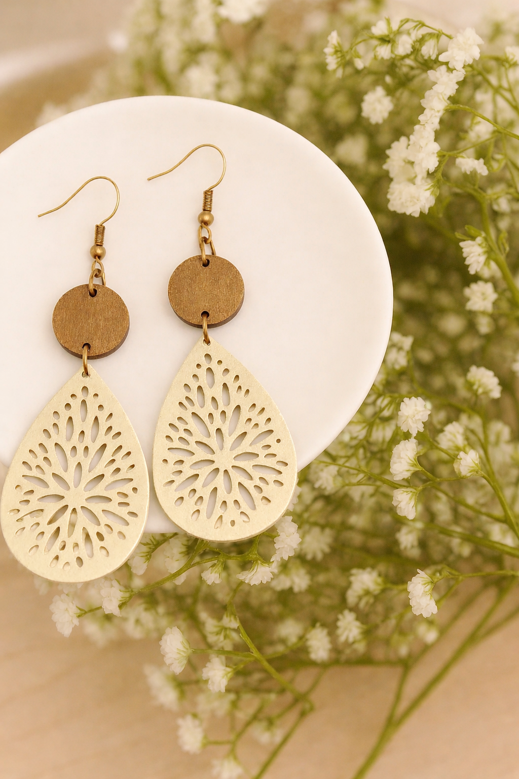 A pair of wooden and metal earrings featuring round brown studs and light gold tear-drop shaped laser-cut floral pendants displayed on a white dish with soft white baby's breath flowers in the background