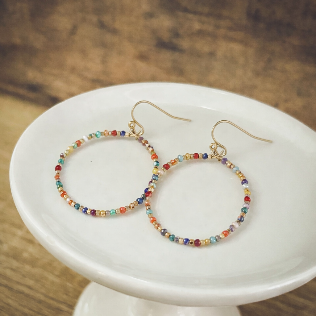 Handmade beaded hoop earrings with gold-toned hooks and vibrant multicolored seed beads displayed on a white ceramic stand against a blurred wooden surface