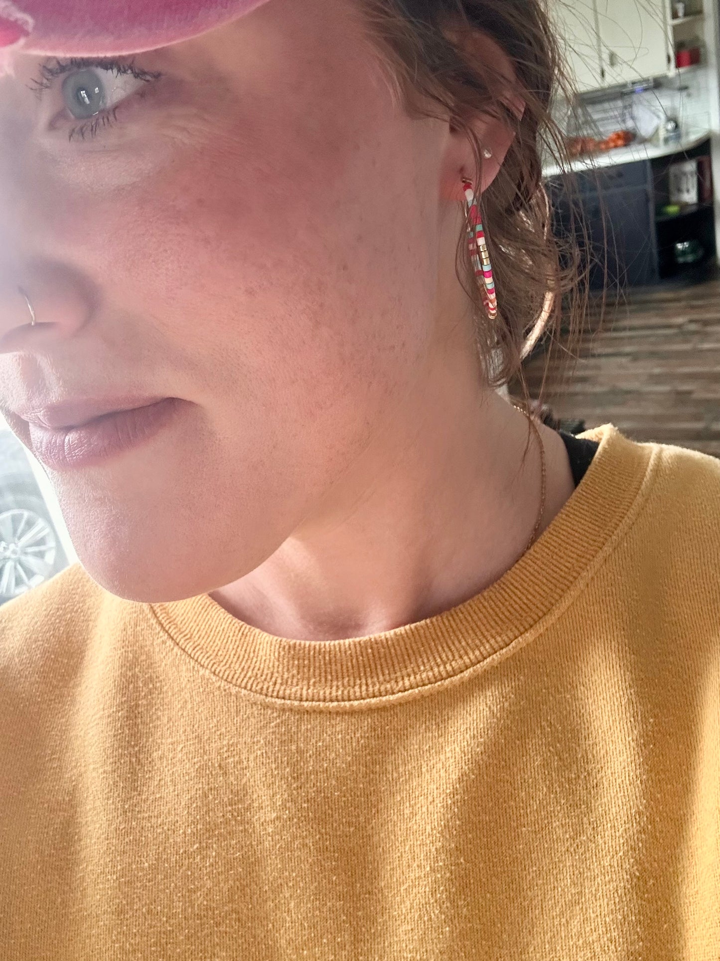 Close-up of a person's face looking upwards, wearing a mustard yellow sweatshirt, a silver nose ring, and colorful beaded dangle earrings against a blurry kitchen background