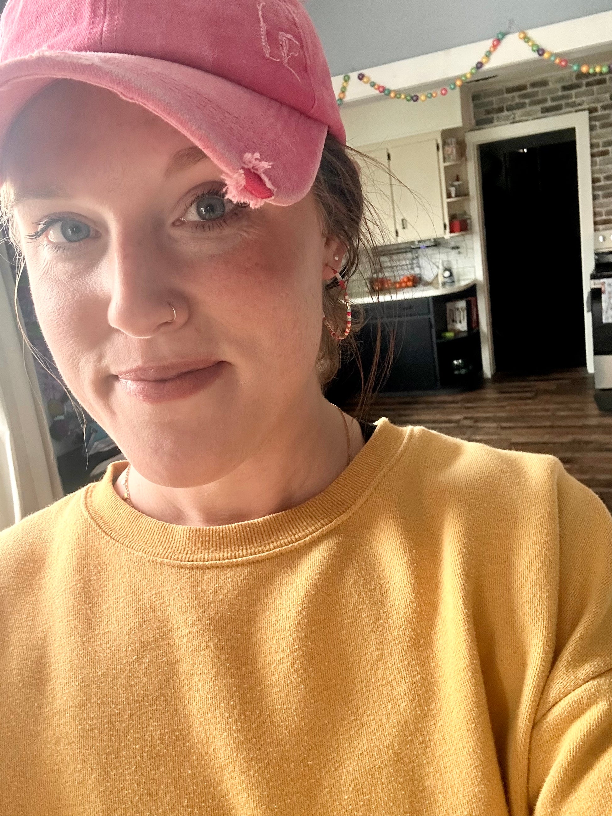 A smiling woman in a distressed pink baseball cap and mustard yellow sweatshirt takes a selfie in a home kitchen with a colorful beaded garland strung across a doorway