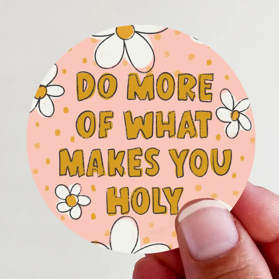 Round pink sticker with orange polka dots and white daisy flowers featuring bold gold text "DO MORE OF WHAT MAKES YOU HOLY" held by a person's fingers