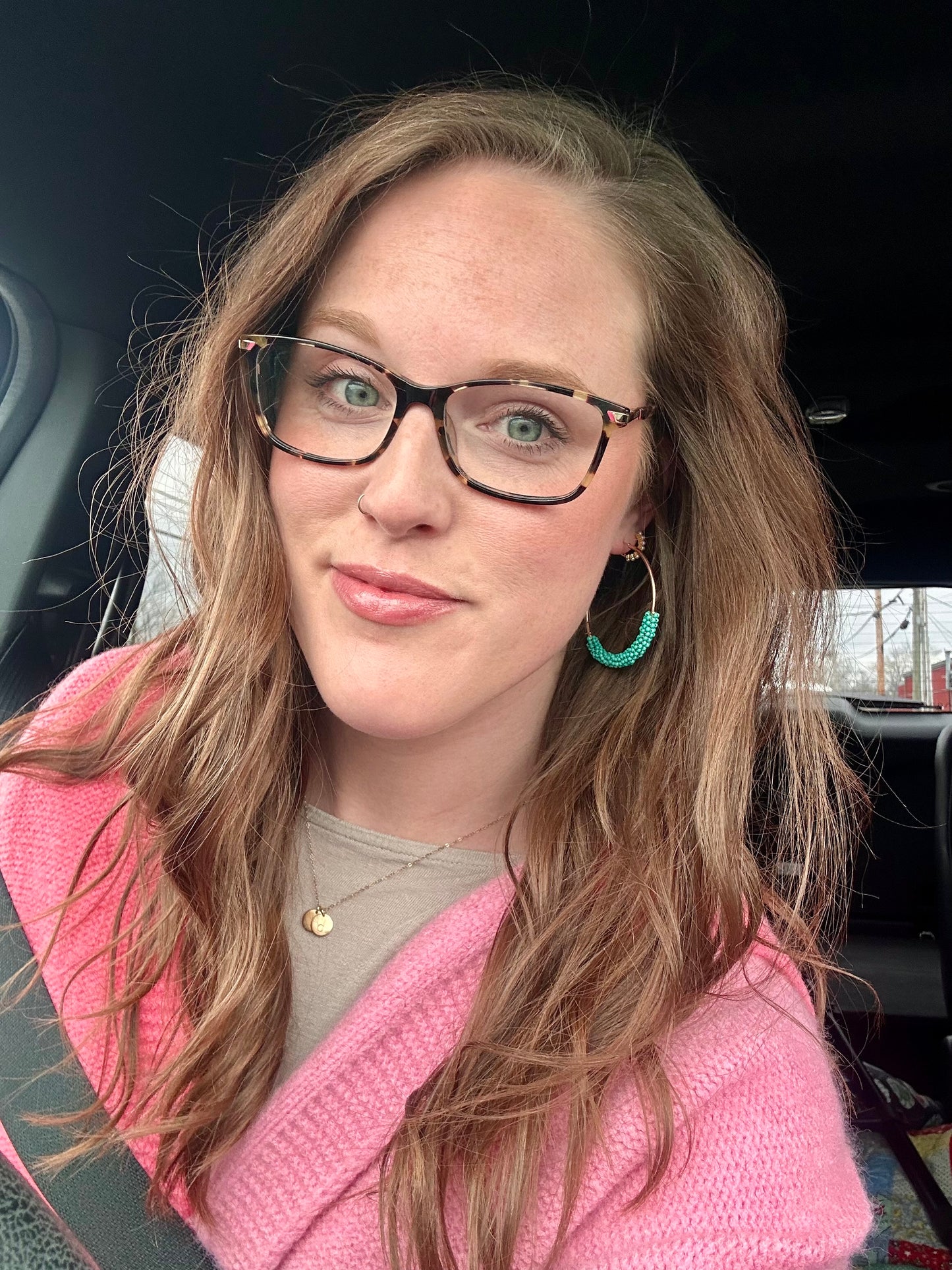 Photograph of a smiling woman with light brown hair and green eyes wearing tortoiseshell eyeglasses a bright pink sweater and turquoise beaded hoop earrings inside a car