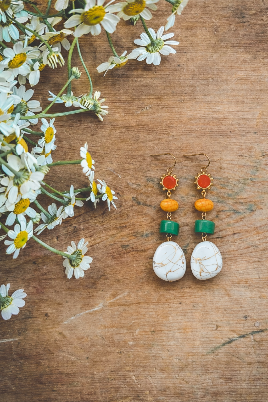 A pair of colorful drop earrings featuring red, golden yellow, green, and white marbled stones with gold veining, displayed on a rustic wooden surface beside a cluster of white daisies