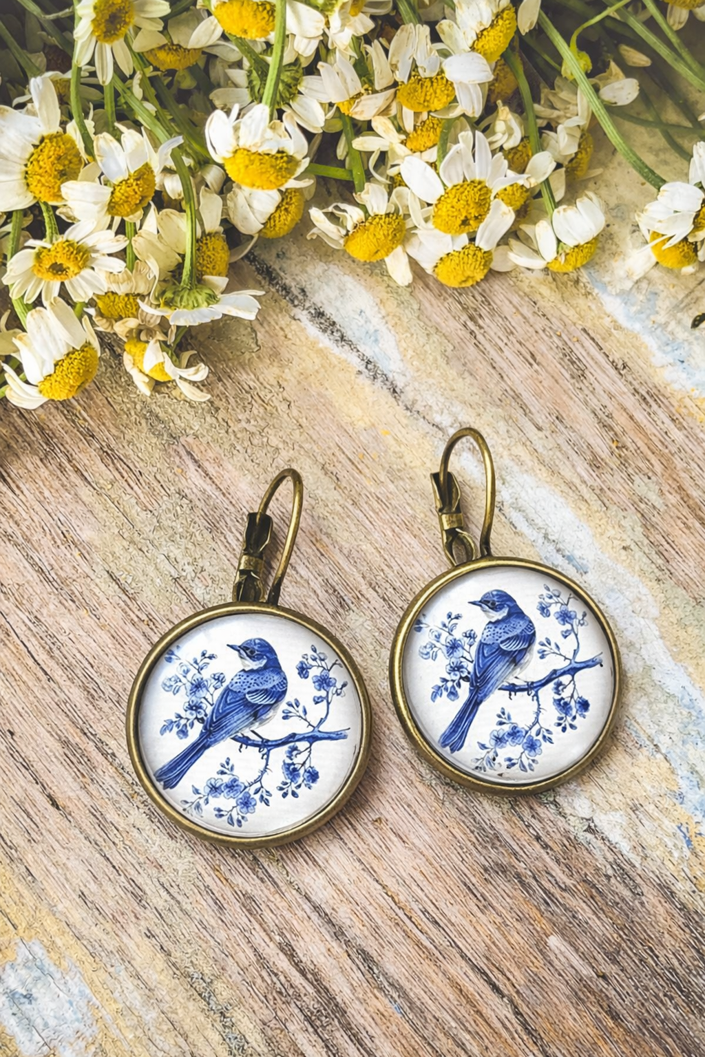 Jewelry: A pair of bronze lever-back earrings featuring round white ceramic cabochons with intricate blue Chinoiserie style birds on branches, displayed on a rustic wooden surface surrounded by white and yellow chamomile flowers