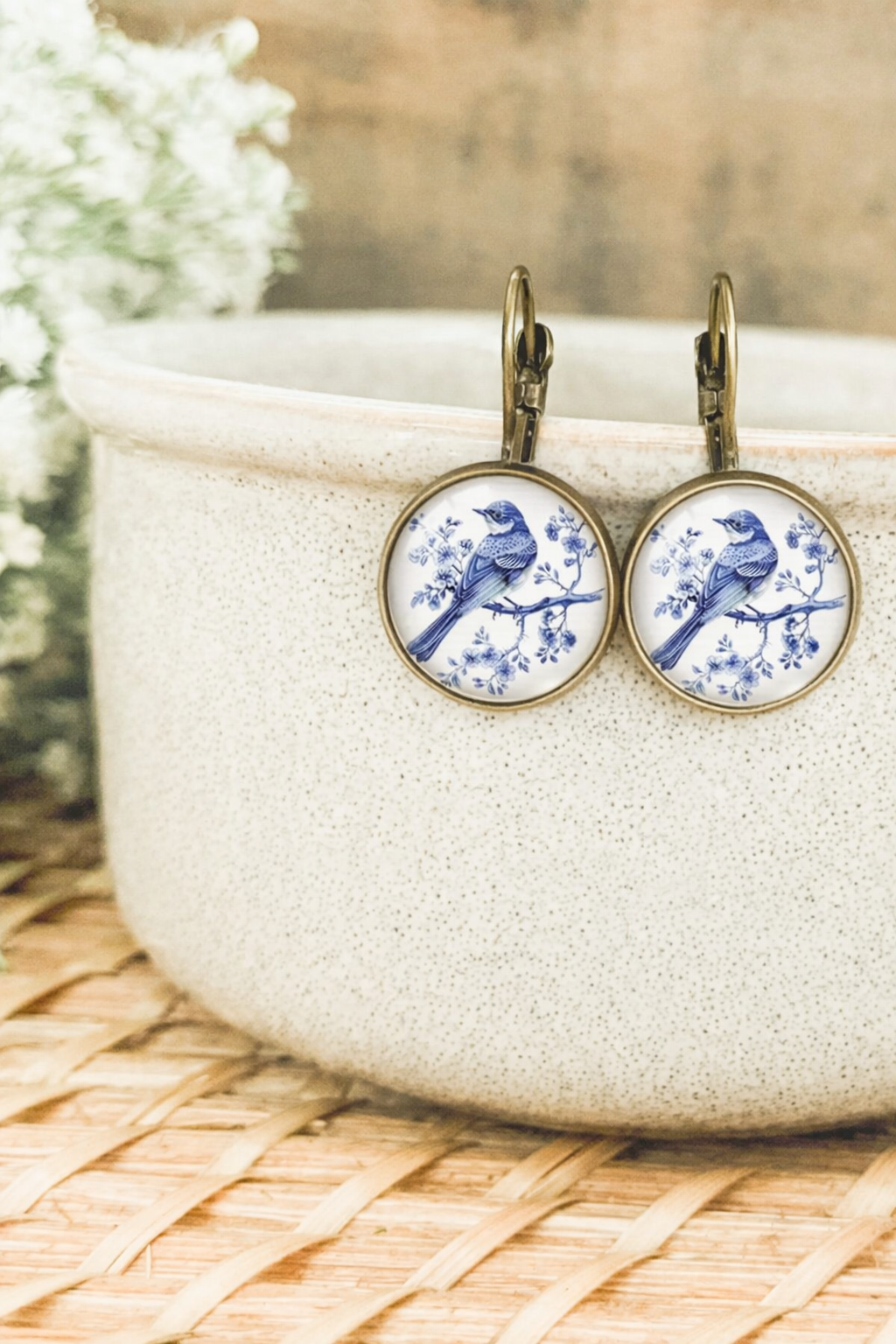 A pair of round antique brass lever-back earrings featuring a delft blue bird perched on a flowering branch illustration, positioned against a speckled ceramic pot and a woven straw mat