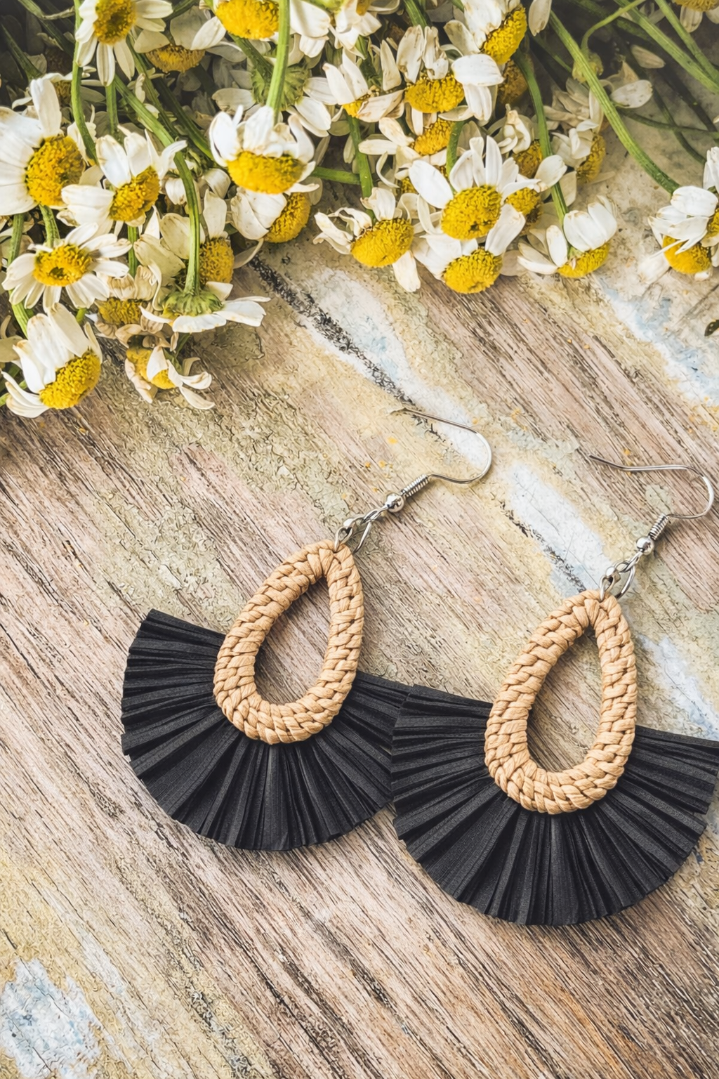 A pair of light tan woven teardrop earrings with black pleated fan details on a rustic wooden surface adorned with white and yellow daisies