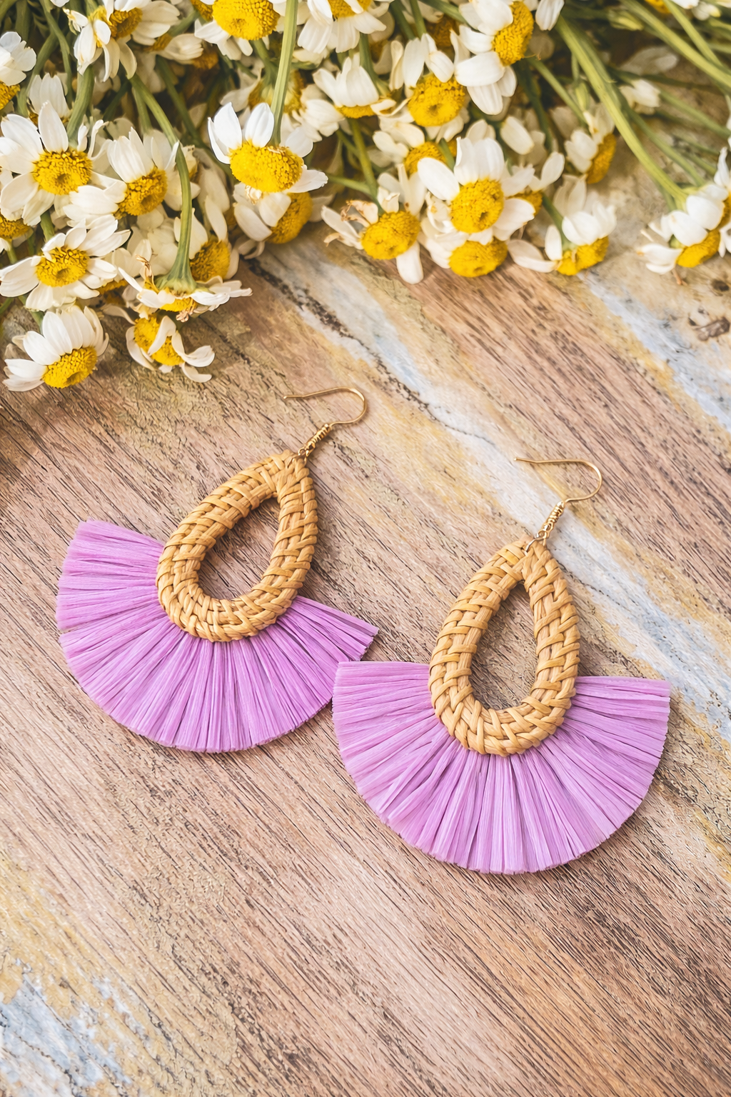 A pair of bohemian earrings featuring woven rattan teardrops and vibrant purple raffia fringe fans with gold-colored hooks, displayed on a rustic wooden surface alongside white and yellow daisies