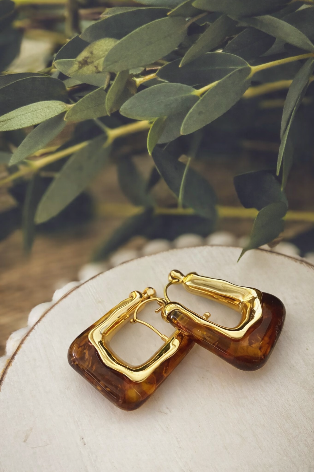 Pair of gold and amber tortoiseshell rectangular hoop earrings on a white wood slice with soft green eucalyptus leaves in background