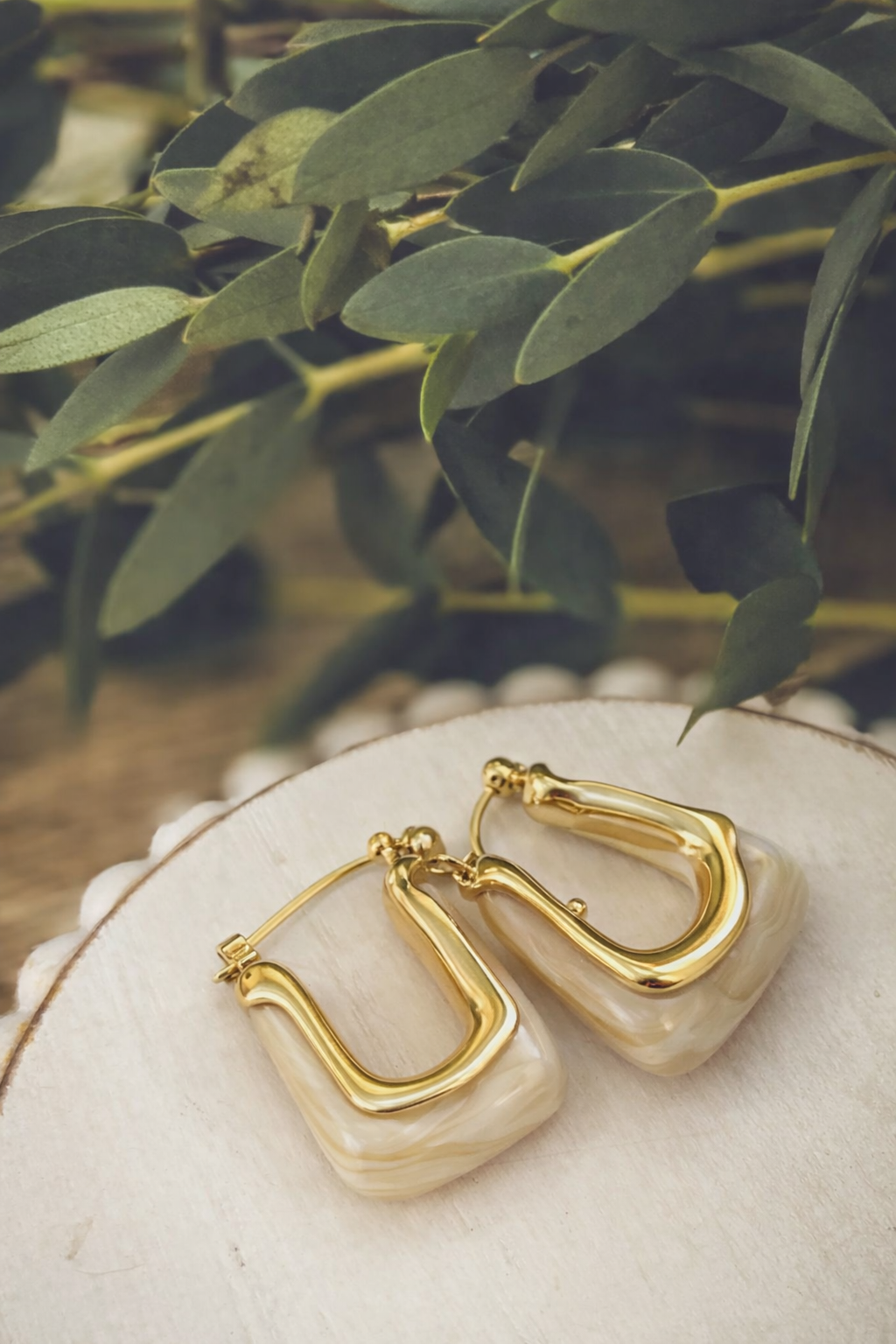 A pair of gold-toned square hoop earrings featuring a cream marbled resin inlay displayed on a white-washed wooden slice with blurred green eucalyptus leaves in the background