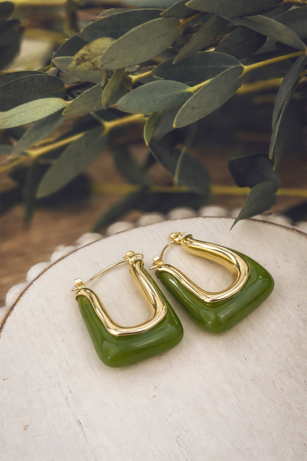 A pair of gold-toned U-shaped hoop earrings featuring forest green rectangular resin inserts displayed on a rustic white wood slice with blurred eucalyptus leaves in the background