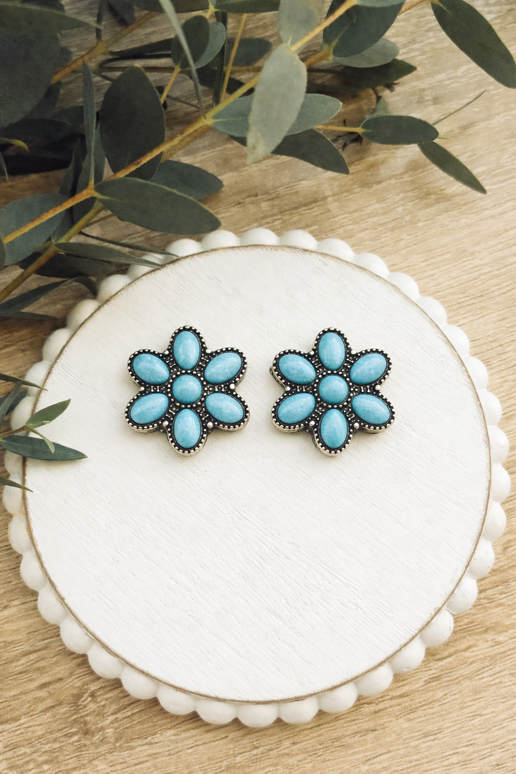 Photograph of a pair of flower-shaped earrings featuring vibrant turquoise stones and detailed silver settings displayed on a white scalloped wooden plate with eucalyptus leaves in the background