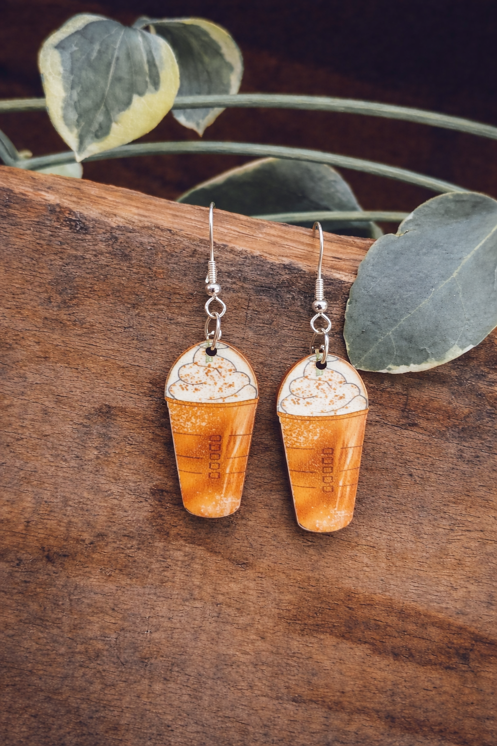Handcrafted earrings, a pair of coffee cup dangle earrings designed like Starbucks Frappuccinos with whipped cream and subtle barcode-like "00000" details, resting on a textured wooden board with variegated green leaves