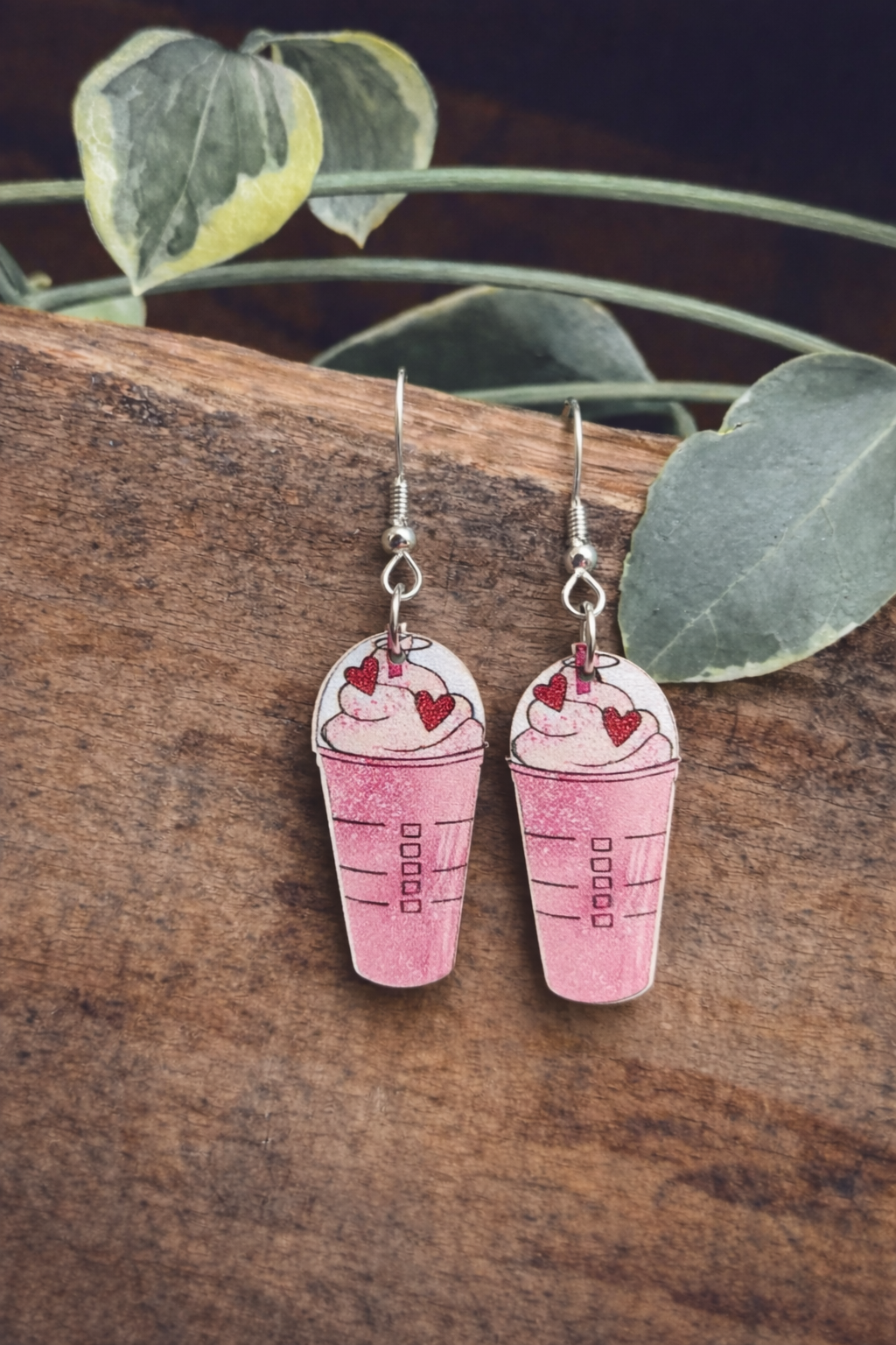 Handcrafted earrings resembling pink Starbucks Frappuccino cups adorned with white whipped cream and red glitter hearts, suspended by silver hooks against a rustic dark wood surface with green variegated leaves