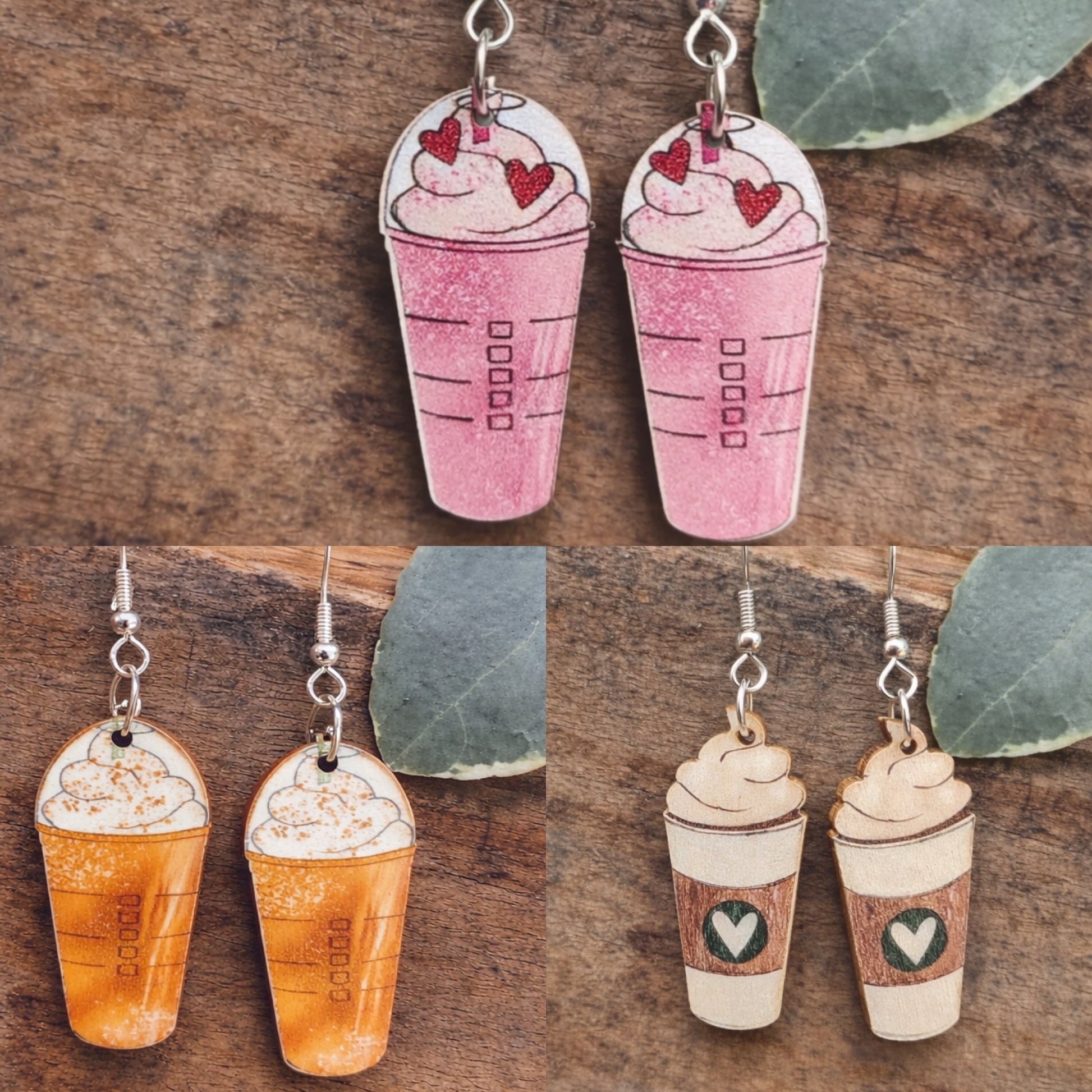 Four pairs of handmade coffee-themed dangle earrings displayed on a rustic wooden surface with green eucalyptus leaves, including pink glitter frappuccinos with red hearts, pumpkin spice lattes with whipped cream, and white coffee cups with green heart logos