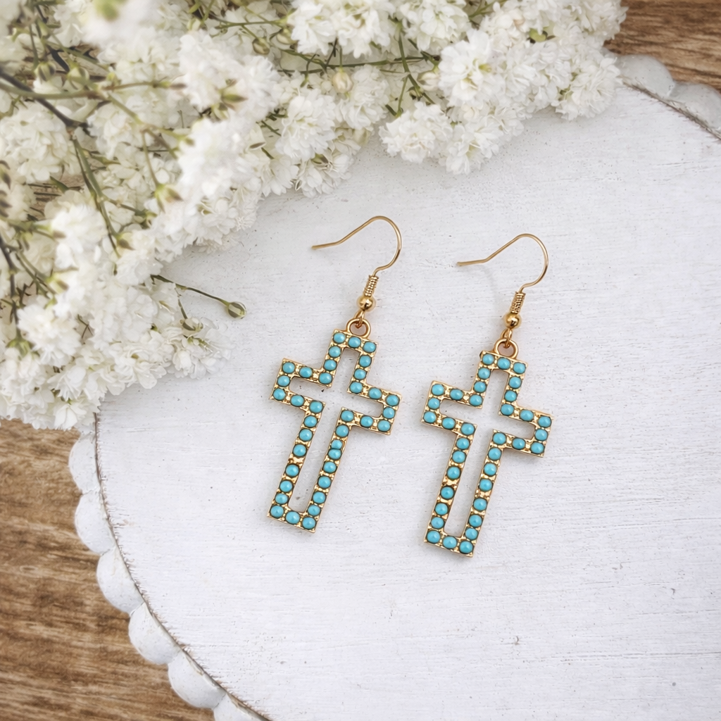 A pair of gold-toned cross earrings with turquoise beaded outlines displayed on a white scalloped tray with delicate white baby's breath flowers in the background
