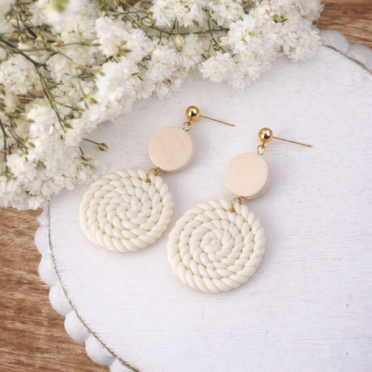 Product photography of a pair of cream coiled rope and light wood dangle earrings with gold-tone hardware on a white scalloped tray surrounded by delicate white baby's breath flowers