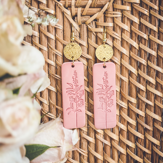 Polymer clay and gold-toned metal dangle earrings, featuring rectangular pink drops with debossed botanical designs and textured gold disc tops, displayed against a woven wicker background with blurred light pink flowers