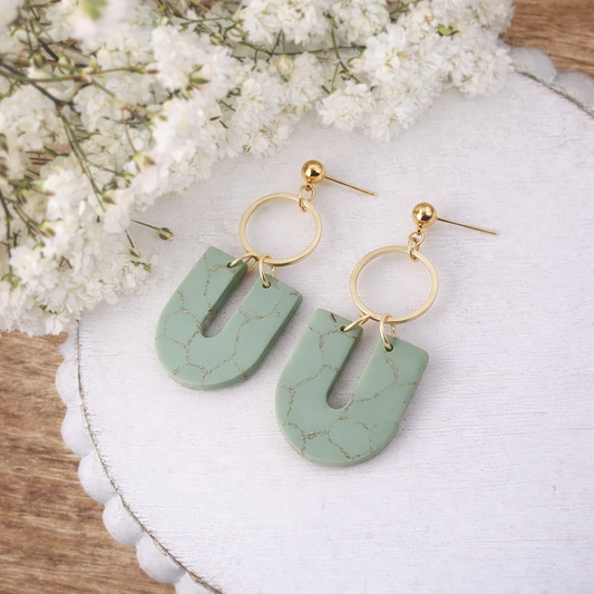 Jewelry a pair of sage green U-shaped earrings featuring gold crackle patterns and gold-toned hoops displayed on a white scalloped tray with delicate white baby's breath flowers in the background