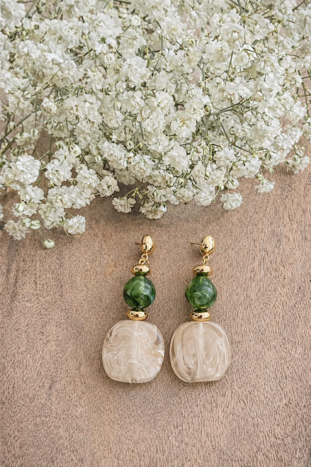 A pair of elegant dangle earrings featuring gold-toned posts, swirled green marbled beads, and opaque cream oval drops, resting on a warm wooden surface beneath a spray of delicate white baby's breath flowers