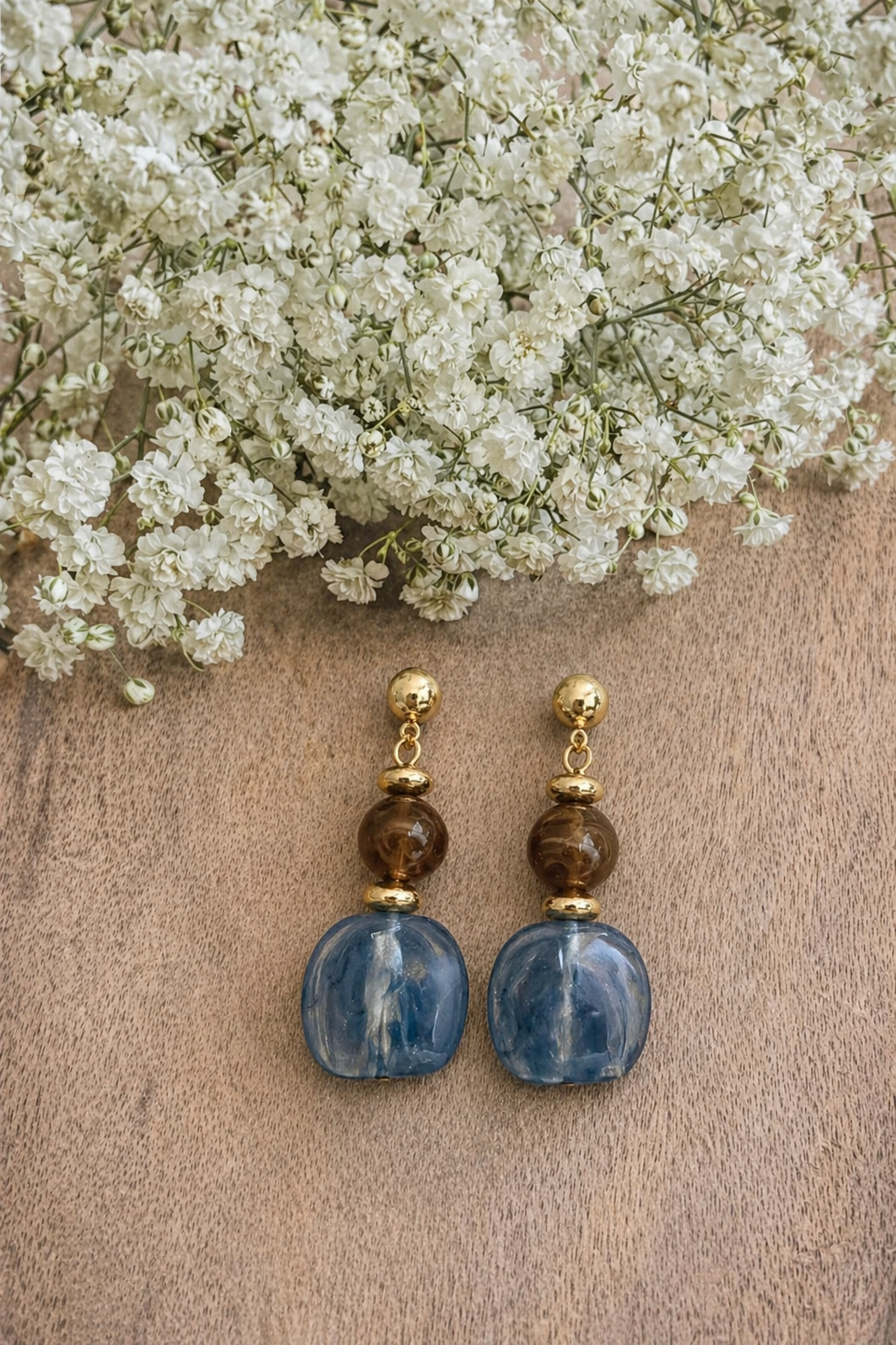 A pair of gold-tone dangle earrings featuring marbled brown and translucent blue rounded beads resting on a natural wood surface under a soft cluster of white baby's breath flowers