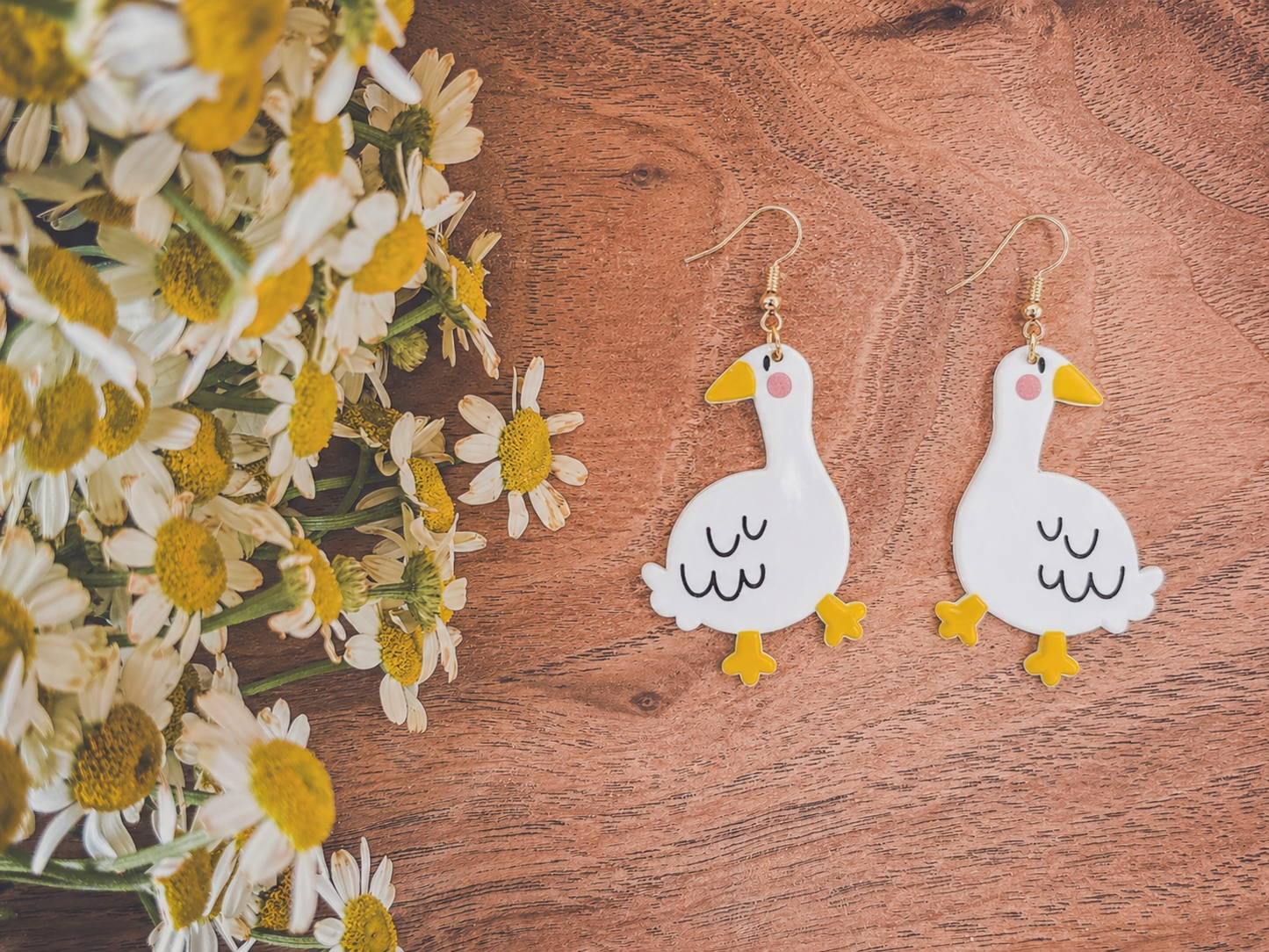 A pair of whimsical white goose earrings with yellow beaks and pink cheeks displayed next to a bunch of fresh white and yellow chamomile flowers on a rustic wooden background