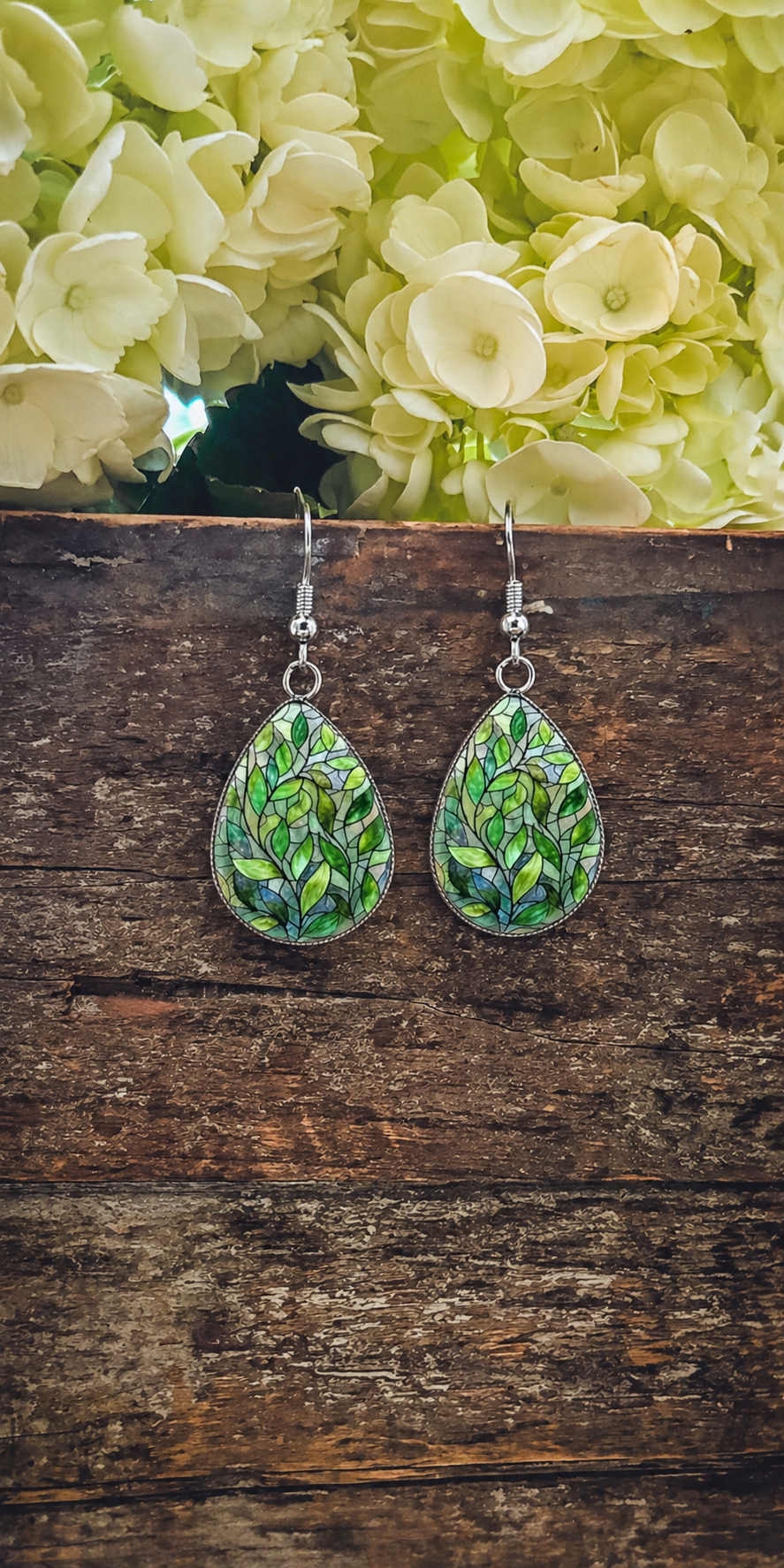 A pair of teardrop-shaped dangle earrings featuring a stained-glass style pattern of various green leaves and blue accents hangs against a rustic dark wooden surface with blurred white and light green hydrangeas blooming above