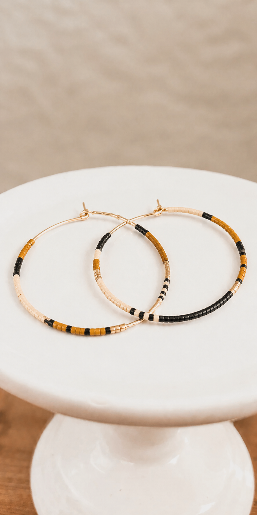 A pair of gold-toned hoop earrings featuring sections of black, cream, and mustard yellow seed beads arranged on a white pedestal