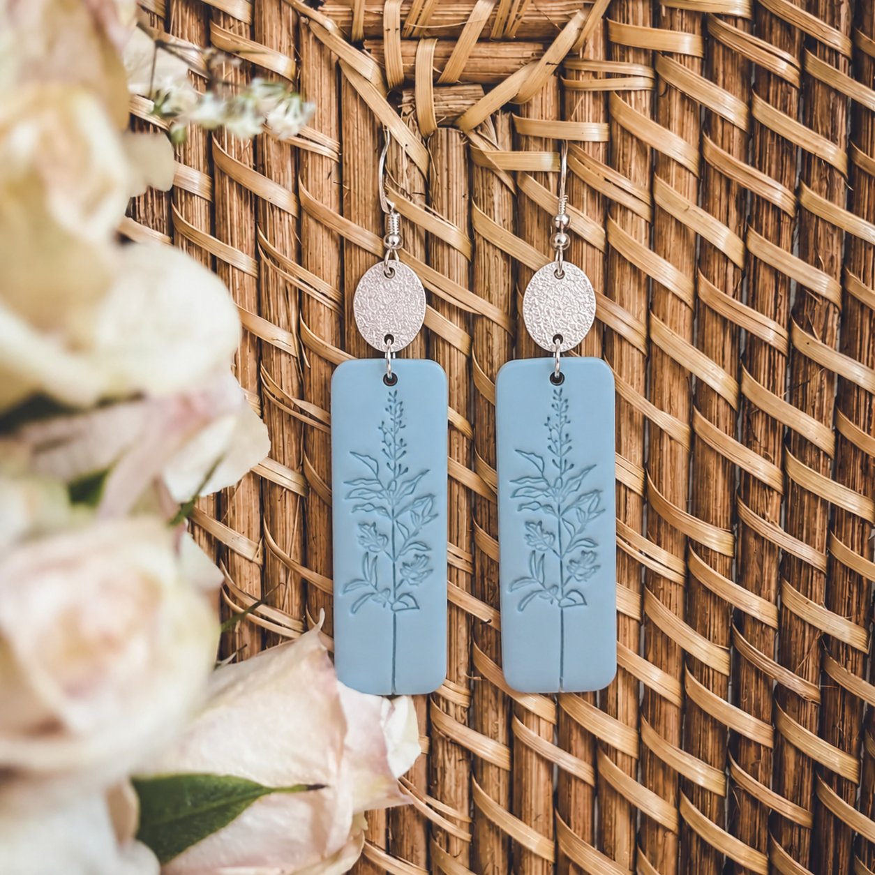 Handcrafted light blue polymer clay rectangular earrings featuring an embossed botanical wildflower design and textured silver ear wires displayed against a woven wicker background with soft focus pale roses