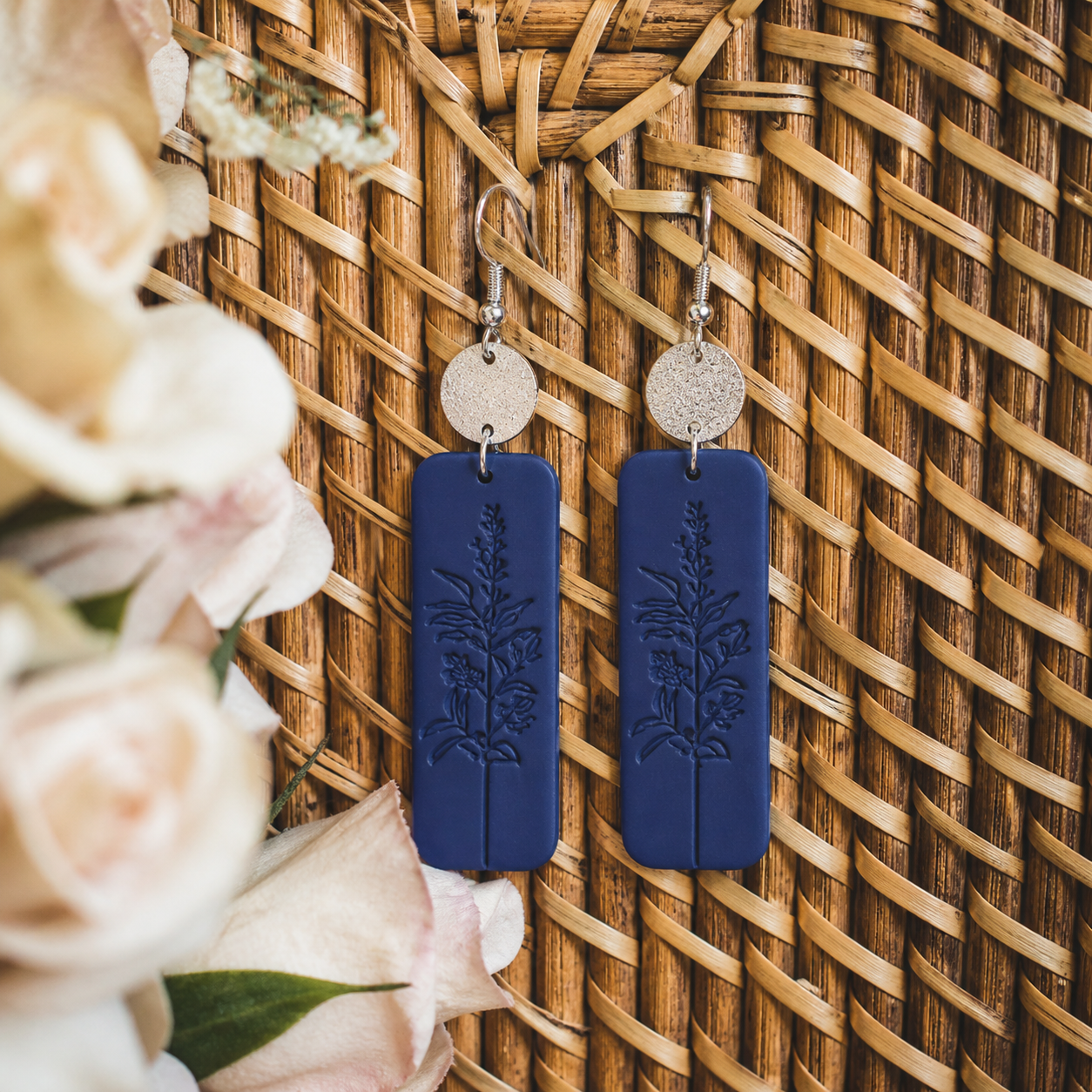Handmade polymer clay earrings, a pair of dark indigo rectangular drops with an embossed botanical design and textured silver studs, displayed on a woven wicker background alongside blurred cream roses