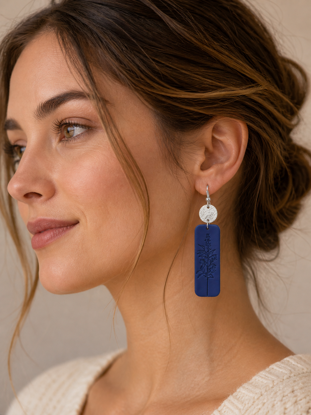 Photograph of a woman's profile with brown hair wearing a cream sweater and a striking dark blue rectangular earring featuring an embossed botanical design and a silver textured disc top