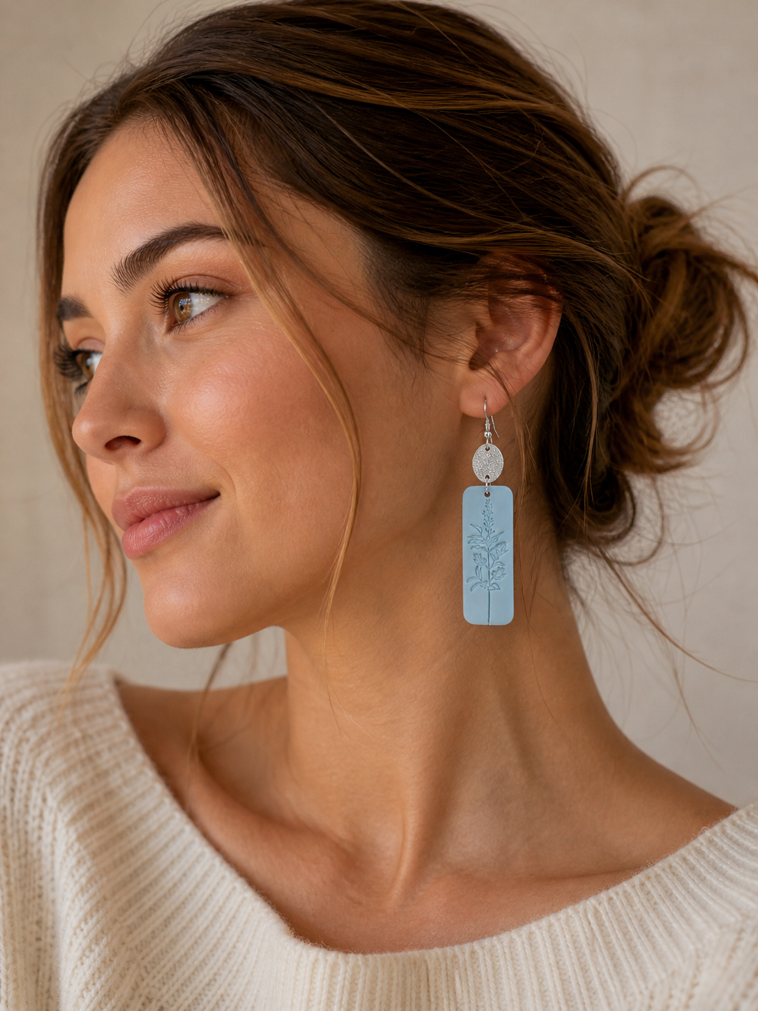 Photograph of a young woman in profile wearing a light blue rectangular dangle earring with a debossed botanical design, a textured silver disc above it, and a cream knit sweater