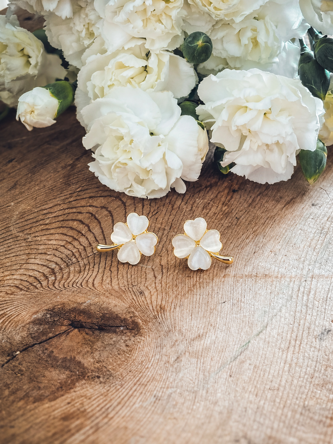 Gold-rimmed mother-of-pearl clover earrings on a rustic wooden surface next to white fluffy carnations with green buds and prominent wood grain