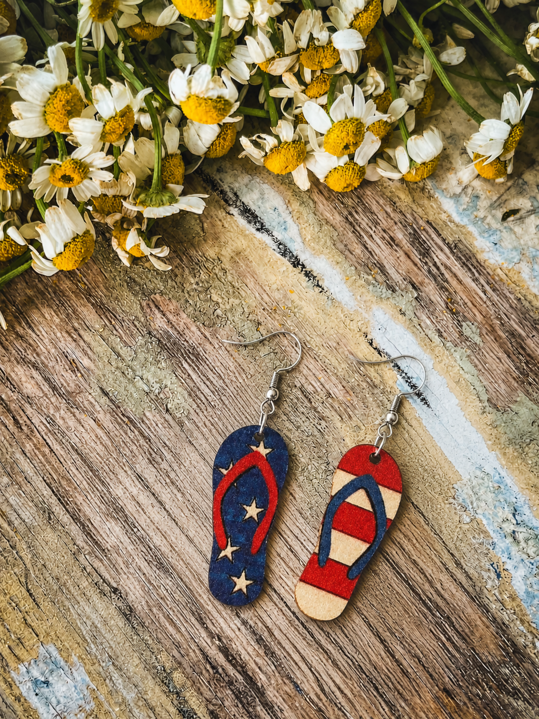 A pair of patriotic flip-flop earrings, one blue with white stars and red straps, the other red and white striped with blue straps, rests on a distressed wooden surface beneath a cluster of white and yellow daisies