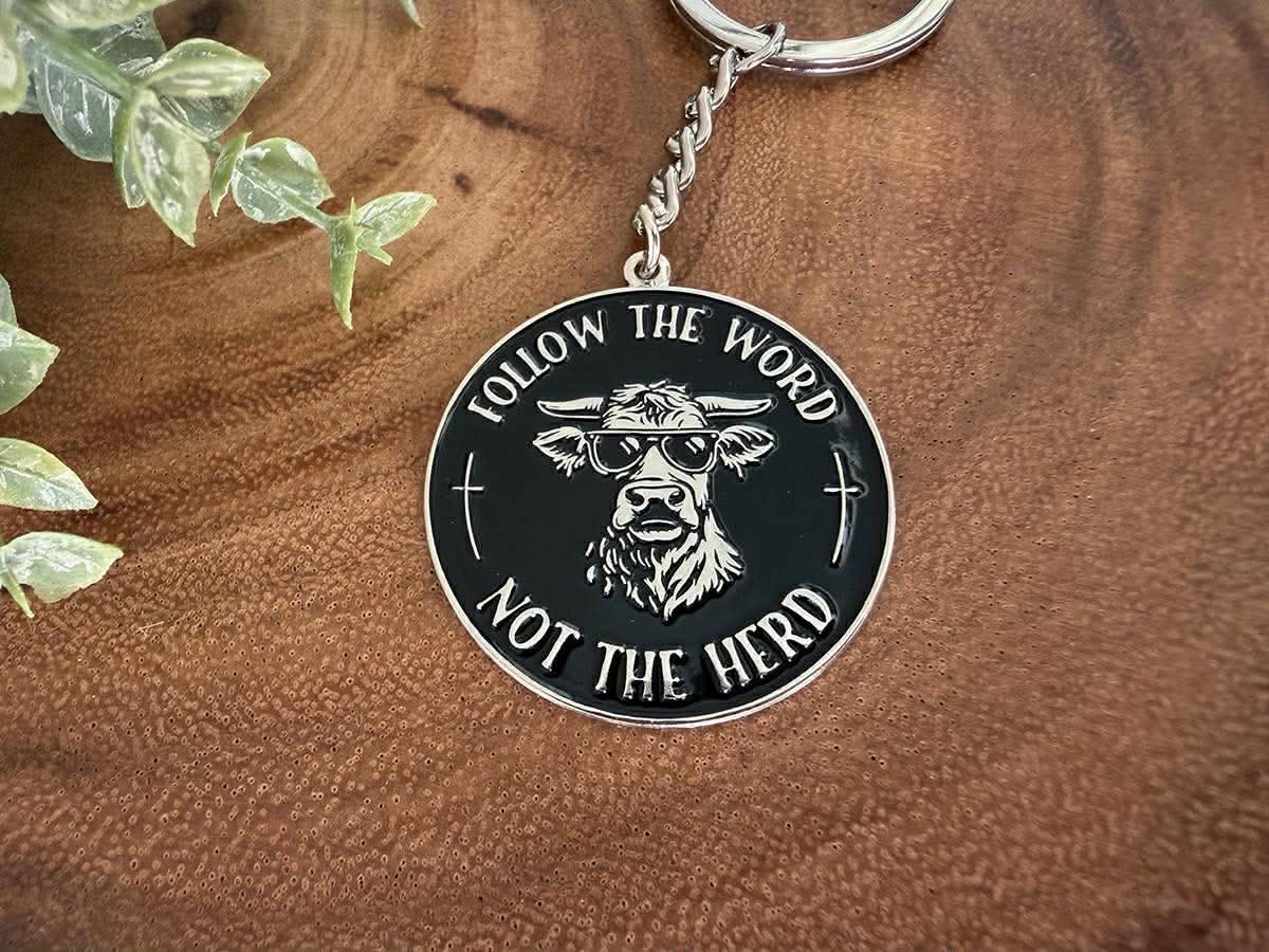 Metal keychain featuring a black and silver round design with a cow in sunglasses and the text FOLLOW THE WORD NOT THE HERD, on a textured brown wood background with green leaves
