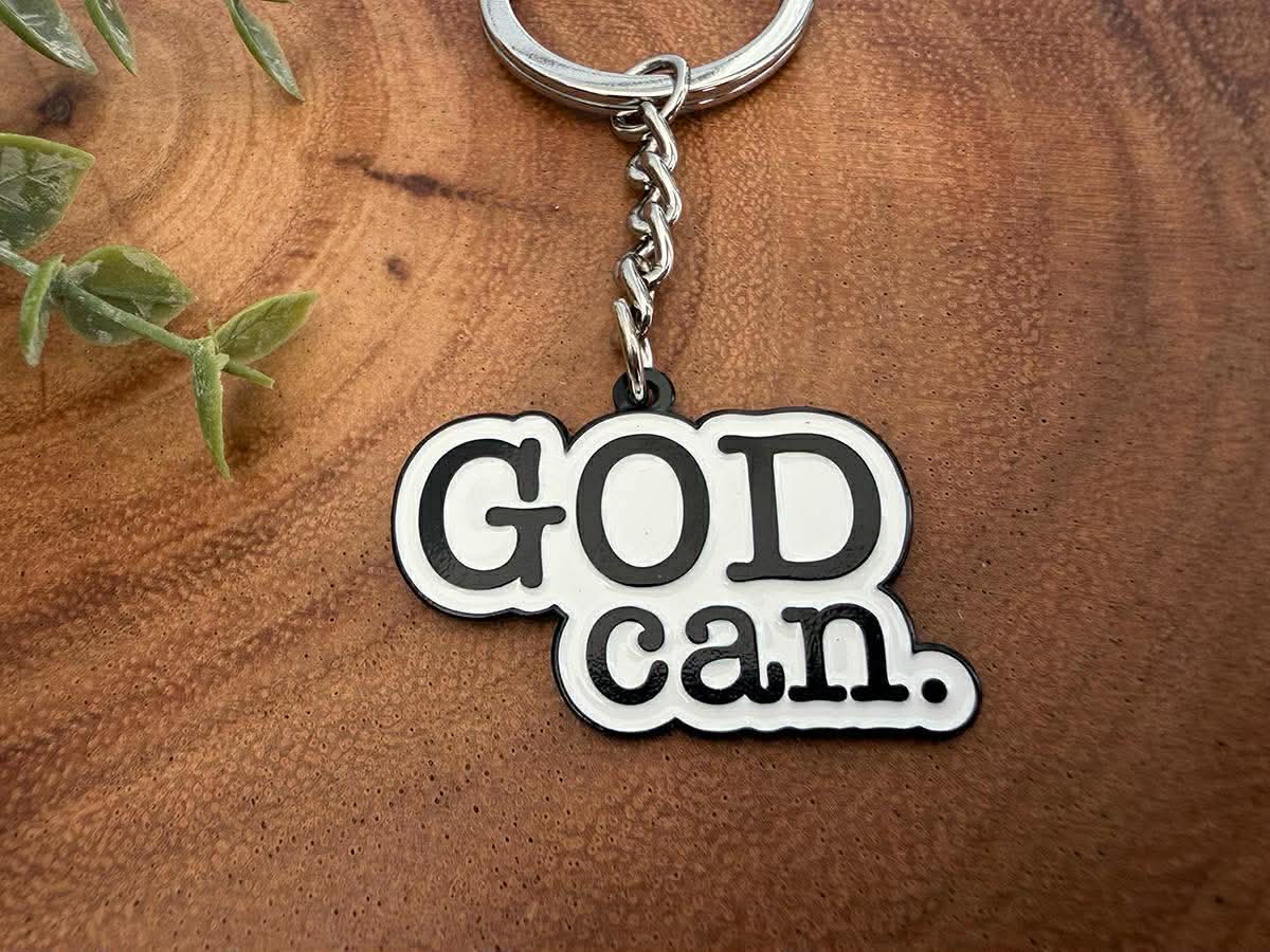 A religious "God can" keychain with a silver ring and chain features bold black text on a white cutout, resting on a textured brown wooden surface next to a blurry green plant sprig
