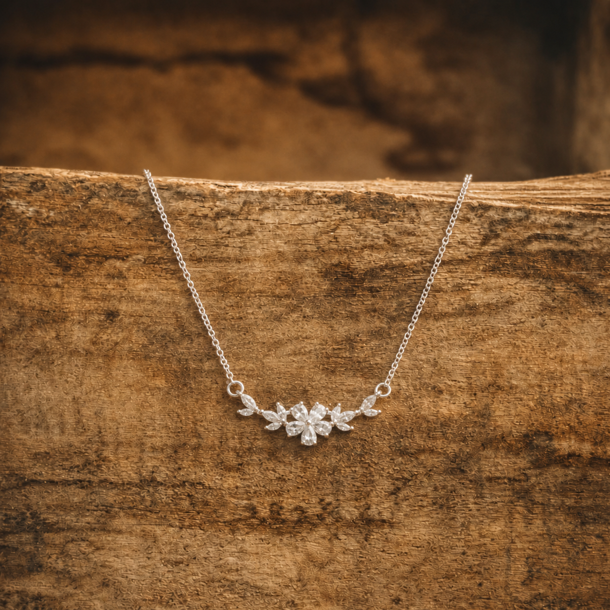 Photograph of a silver chain necklace with a sparkling clear gemstone floral cluster pendant resting on a rustic wooden surface