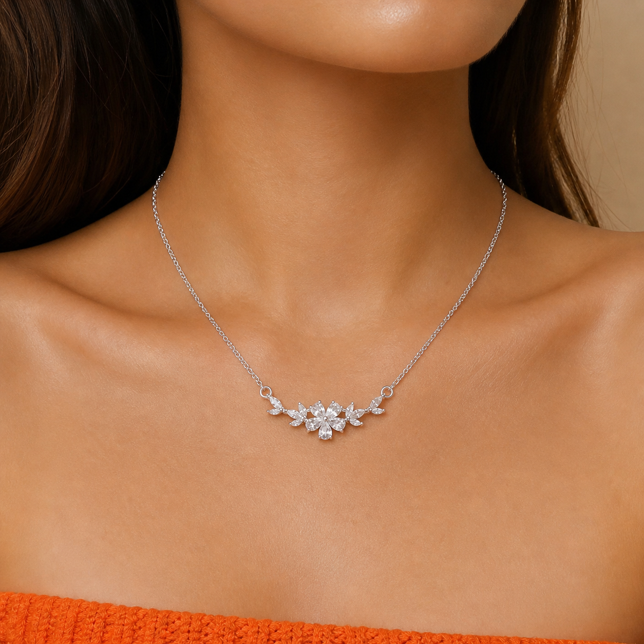 Photograph of a woman's décolletage displaying a delicate silver chain necklace with a floral cluster pendant adorned with sparkling marquise and round cubic zirconia stones, resting above an orange knitted garment