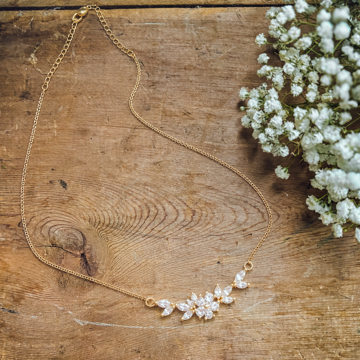 Photograph of a delicate gold-toned necklace featuring sparkling clear cubic zirconia floral clusters laid on a rustic wooden surface next to a sprig of white baby's breath flowers