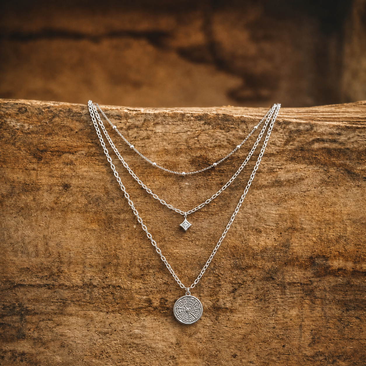 Three silver layered necklaces featuring a delicate beaded chain a small diamond rhombus pendant and an intricately engraved circular pendant all displayed on a rustic brown wooden background