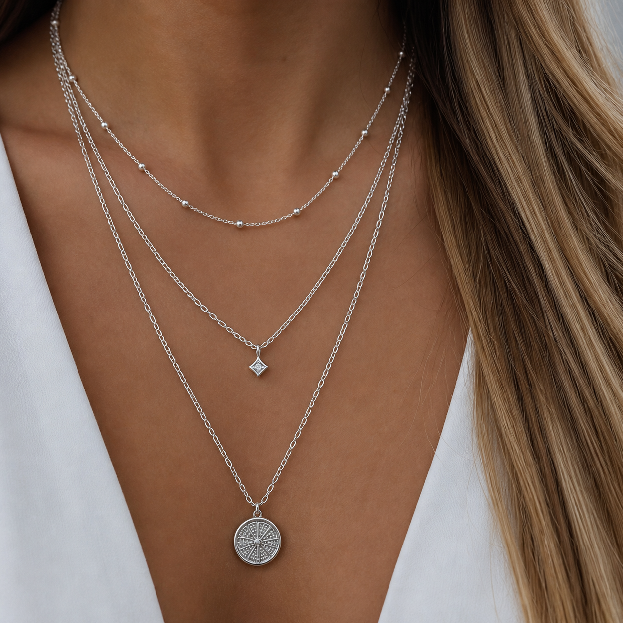 Photograph of a woman's décolletage featuring three layered sterling silver necklaces including a delicate beaded chain, a small square diamond charm, and a round compass rose coin pendant, worn with a white shirt and blonde hair