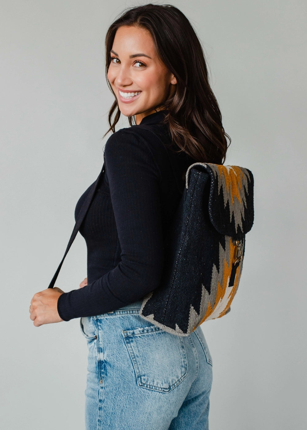 Smiling woman with dark wavy hair wearing a black ribbed long-sleeve top and light wash jeans models a navy gold grey and white chevron woven backpack against a plain studio background