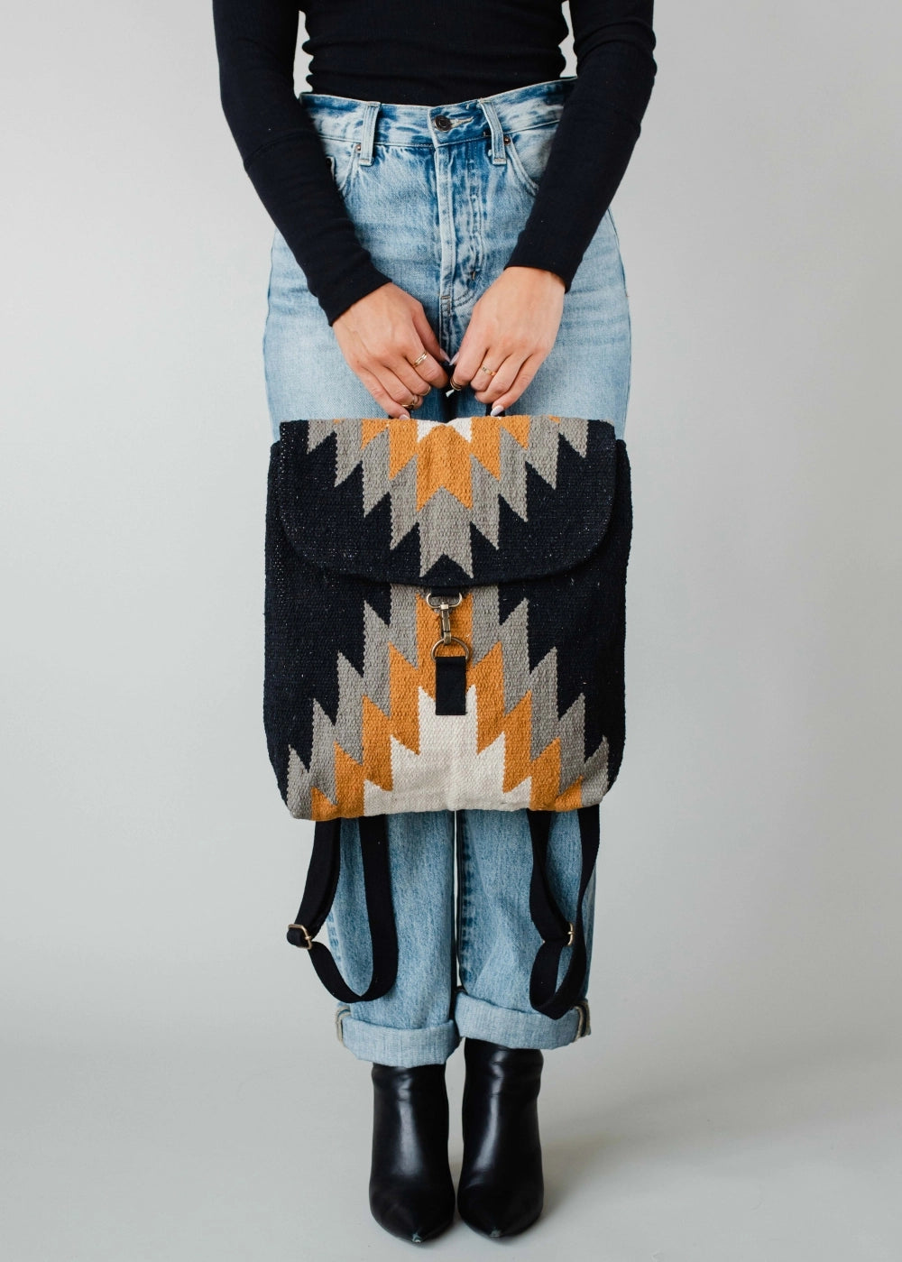 Photograph of a person in cuffed light wash jeans and black ankle boots holding a textured backpack featuring a striking geometric zigzag pattern in black, grey, white, and golden yellow against a plain studio background