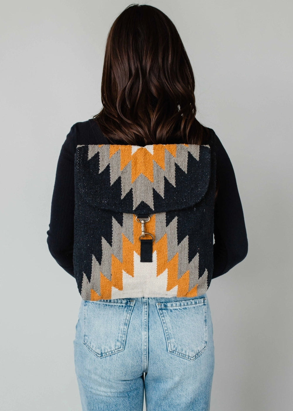 Photograph of a person seen from behind wearing a black long-sleeve shirt, light wash blue jeans, and a textured dark blue backpack with a striking orange, grey, and white zig-zag pattern featuring a silver hook clasp on a plain studio background