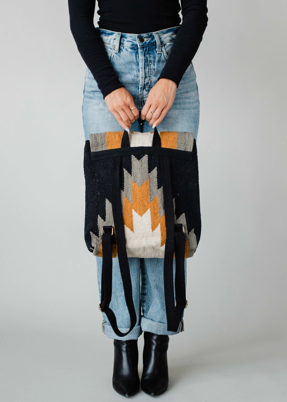 A person wears cuffed blue jeans and black ankle boots while holding a handwoven geometric patterned backpack in black, grey, cream, and mustard yellow against a light studio background