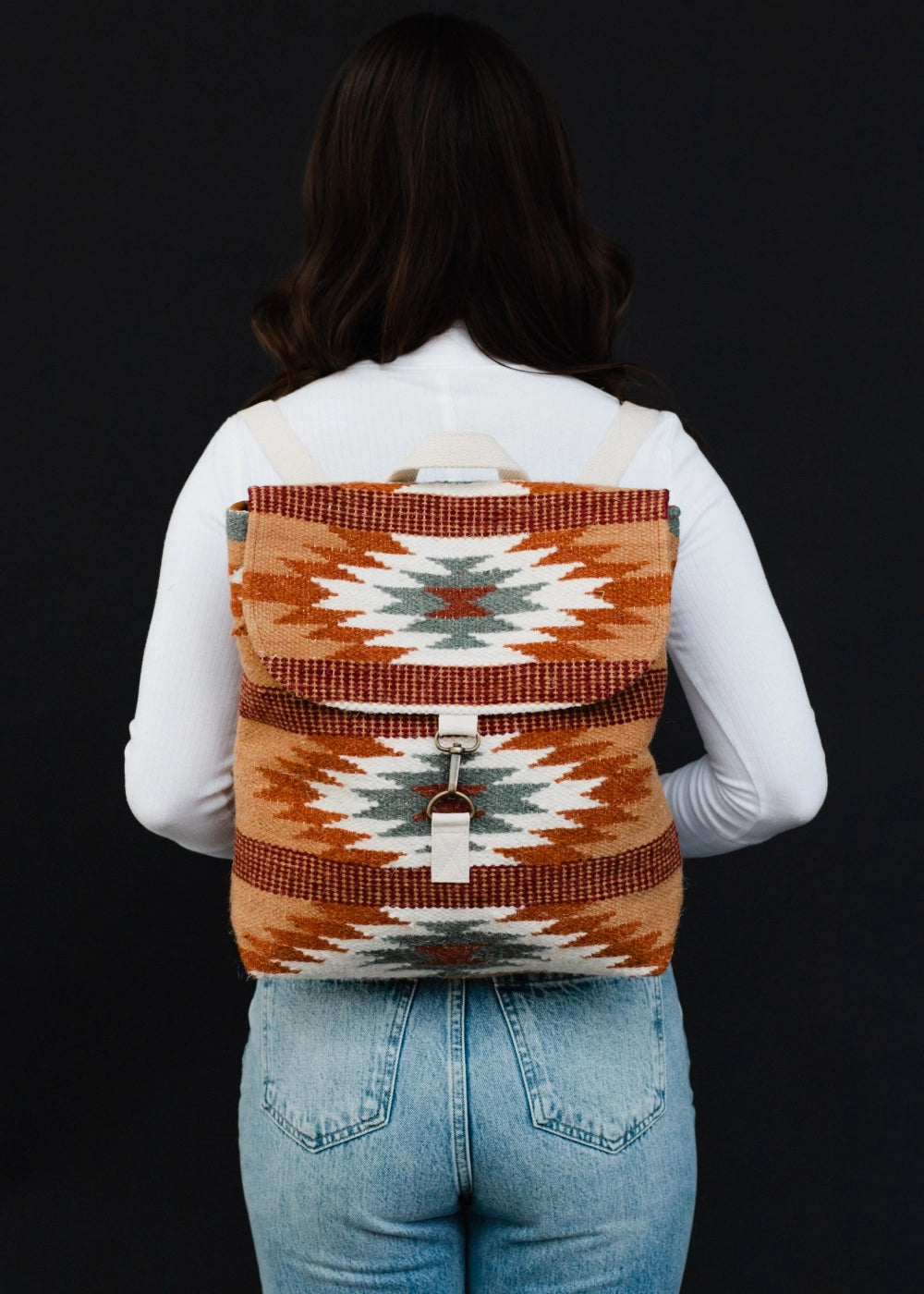 Photograph of a woman from behind wearing a Southwestern geometric patterned backpack in rust orange teal and white with white canvas straps and a metal hook closure over a white long-sleeve shirt and blue jeans against a black studio background