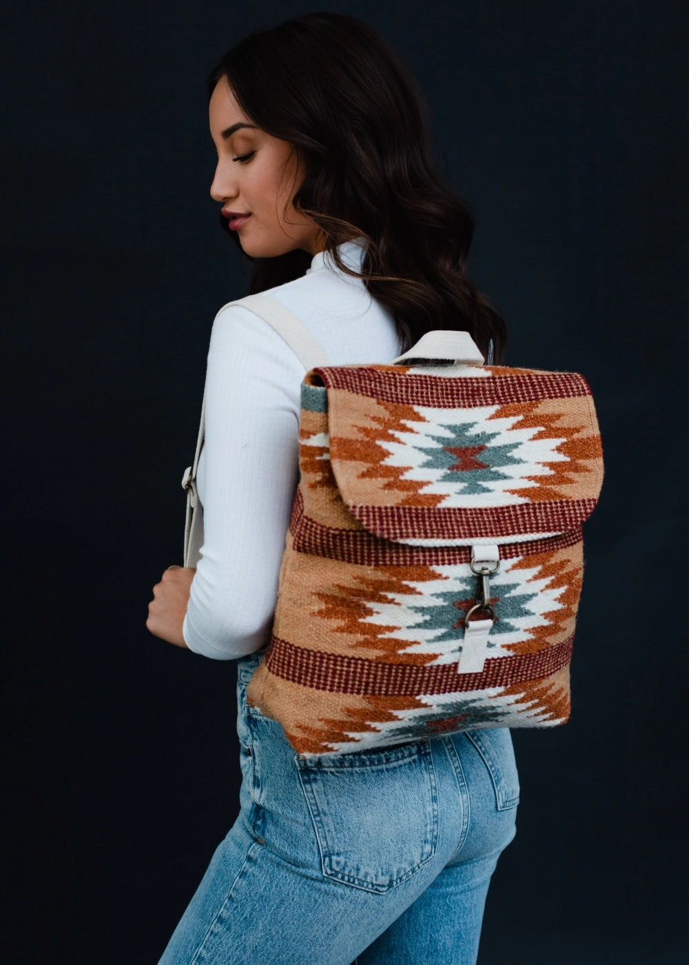 A woman wears a vibrant woven backpack featuring a Southwestern pattern in tan, rust, white, and teal, styled with a white ribbed turtleneck and blue jeans against a dark background